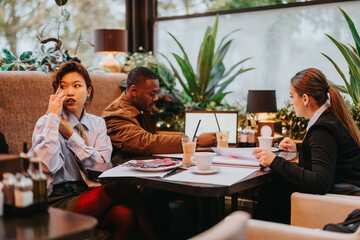 Three business people of different backgrounds engaged in teamwork at a modern cafe, discussing projects, reviewing documents, and working together, all in a dynamic and comfortable setting.