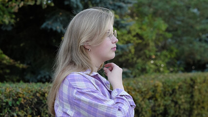 A teenage girl with glasses stands in a green park, looking away with a reflective expression. The...