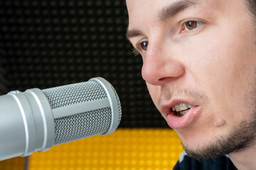 Young man recording voice in professional podcast studio with acoustic background and close-up of...