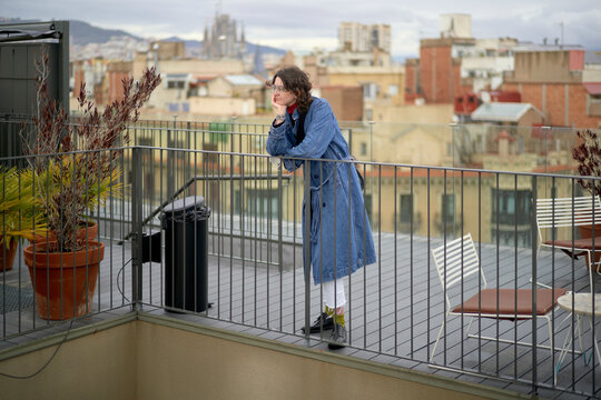 Woman in blue coat leaning on rooftop railing overlooking cityscape with cloudy sky. Brussels