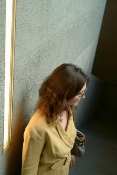 Woman in beige coat standing by a concrete wall with soft lighting.