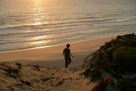A young boy walks down a sandy dune toward the ocean at sunset, with waves gently crashing. Portugal - Powered by Adobe