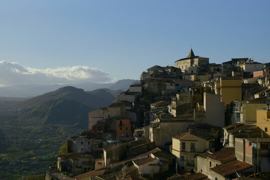 Sunlit hillside town with densely packed houses and distant rolling hills under a clear sky. Sicily, Italy