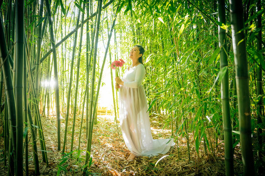 Pregant woman in a white dress holding flowers, standing among tall bamboo stalks. WA, USA