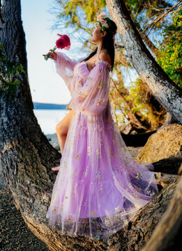 Pregant woman in a pink floral dress stands on tree branch by the sea with a flower. WA, USA