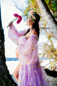 Pregant woman in a pink floral dress stands on tree branch by the sea with a flower. WA, USA