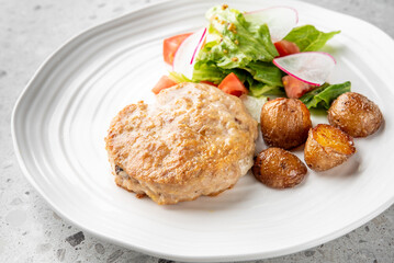 A delicious meal featuring a juicy grilled chicken patty, crispy roasted baby potatoes, and a fresh salad with lettuce, radish slices, and diced tomatoes, served on a white plate.