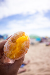 Woman’s hand holding a Bola de Berlim pastry with Portuguese sandy beach in the background, capturing a perfect blend of local snack and coastal scenery.
