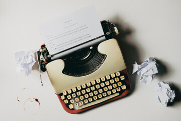 A vintage typewriter is placed on a table with coffee cup rings, and a sheet of paper is inserted. Crumpled pieces of paper lie nearby. The text is an imaginary script written by an imaginary writer. 