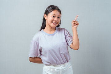 Smiling young Asian girl in a pastel t-shirt pointing upward with one finger while looking at the...