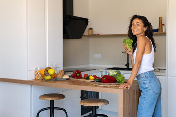 Happy young woman in a white tank top smiling while holding a fresh green lettuce in her hand. Healthy lifestyle concept with vegetables and fruits on a kitchen counter.