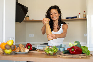 Young woman preparing and seasoning a fresh vegetable salad in a modern kitchen. Full lifestyle...