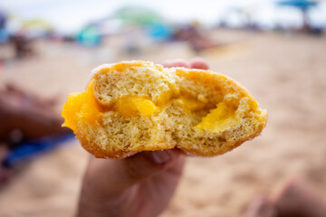 Woman&rsquo;s hand holding a half-eaten Berliner doughnut on a Portuguese beach, with golden sand in the background. A simple summer moment captured close-up.