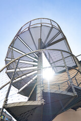 Spiral iron staircase with sunlight streaming through. Dramatic shadows and industrial elegance. Play of light and geometry in modern architectural detail.


