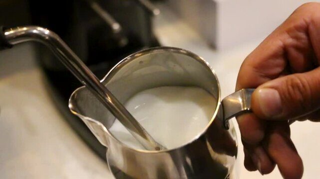 Steam wand heats cold milk. Velvety foam forms in a steel pitcher. The steam wand cuts into the milk. Microfoam texture develops. Bubbles swirl in the liquid. Soft caf&eacute; lighting highlights metal