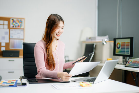 Young graphic designer using pen tablet and laptop at creative desk in modern office. Surrounded by color palettes