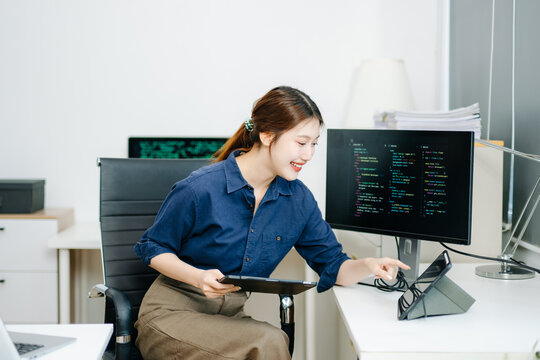 Young female programmer working with tablet and screen displaying code. Modern tech office setup, great for software, coding, IT