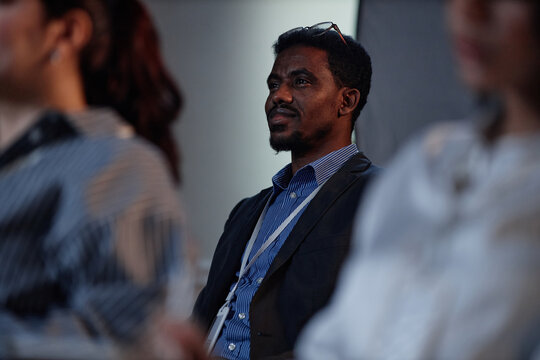 Medium shot of smiling Black male manager wearing conference participants badge sitting in audience space attending executive leadership program meeting in auditorium