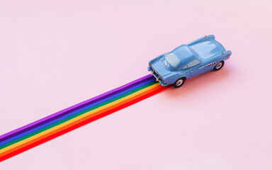 Blue toy car, rainbow ribbon flag trail road, pink background. June gay parade, proud month, coming out day, human rights and tolerance concept. Minimal pop art composition. Flat lay, top view