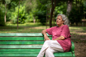 Thoughtful worried indian senior aged woman pensively deep thinking while sitting on bench at park...