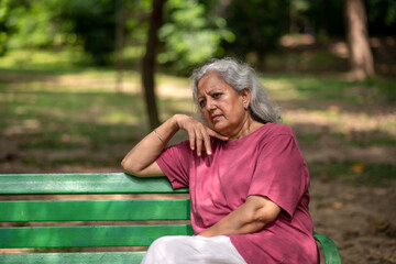 Thoughtful worried indian senior aged woman pensively deep thinking while sitting on bench at park...