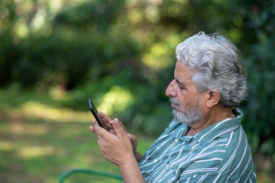 Indian senior man using and text messaging on mobile while standing at park. Indian Senior man using digital technology at outdoors