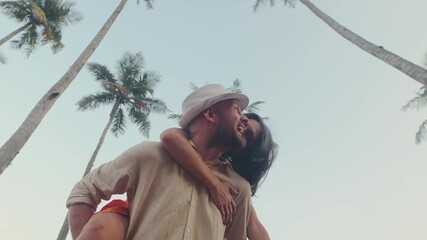 Carefree caucasian happy couple enjoying moment being playful piggyback ride during honeymoon vacation, laughing and sharing joyful moments outdoors in beautiful tropical palm tree blue background - Powered by Adobe