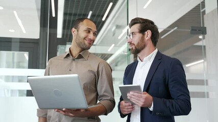 Two smiling businessmen communicate standing in business office. Male colleagues talking during break using laptop. Professionals exchanging ideas for project development. Teamwork and collaboration - Powered by Adobe