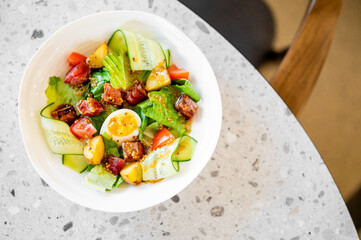 Fresh salad in white bowl on a terrazzo table, featuring mixed greens, cherry tomatoes, cucumber ribbons, diced potatoes, seared tuna, and halved soft-boiled egg, garnished with mustard seed dressing