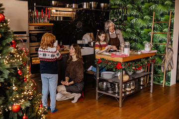 A mother sits and talks to her son who is standing next to her, while a grandmother and granddaughter are cooking at the table in the kitchen