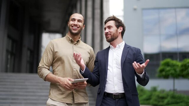 Two happy businessmen communicate while walking outside near office building. Male colleagues have friendly conversation, talking, smiling, laughing. Teamwork, communication and business collaboration