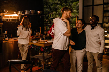 Male and female guests standing and talking to a man walking past them, in the kitchen