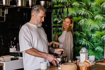 A man is standing at a table and holding a whisk in a bowl while a woman standing next to him is...