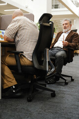 A male worker sits on a chair and holds a pencil while looking at a male worker sitting next to him at a desk