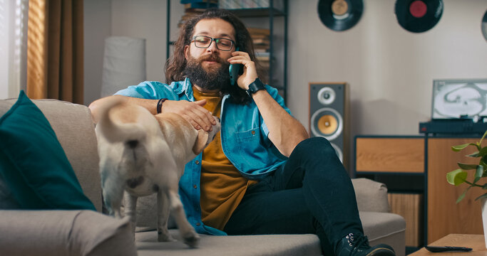 A man sits comfortably on the couch after work, answers his phone and strokes his dog while chatting with a friend.