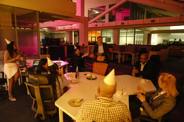 A female employee stands and leans on a table while a group of six employees sit next to her, they applaud and look at a male employee standing next to them, at a party