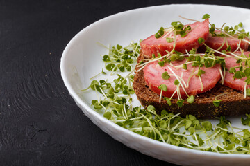 Sandwich with bread and sausage slices, decorated with homegrown fresh harvested broccoli cabbage microgreen sprouts on white plate. Close up