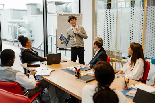 Male intern standing and holding a clipboard while talking to a group of five employees sitting in front of him at a table in an office