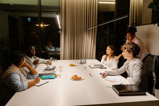 A female employee stands at a table with a male intern and a group of three employees sitting behind it, they are talking, in the office in the evening