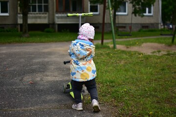 Baby girl pushing green scooter near old soviet apartment houses in cold weather spring. Toddler...