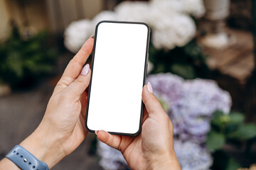 Mockup woman's hand holding using smart phone with white blank screen POV surrounded by soft floral background, mobile app design and advertising, online marketing	