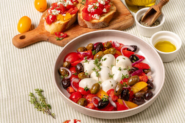 Caprese  salad with colorful tomatoes, mozzarella balls, and a variety of olives, garnished with herbs, served in a bowl on a textured tablecloth with olive oil and bread	