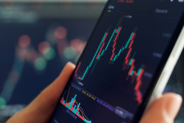 A person analyzes stock trends on a smartphone, studying candlestick charts and trading data, with market indicators blurred behind