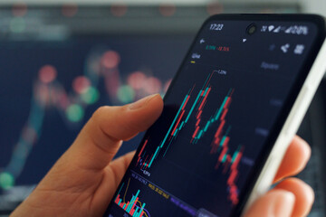 A person analyzes stock market data on a smartphone with colorful graphs displayed, set against a financial trading background