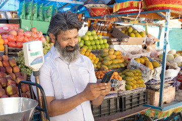 Indian shopkeeper man selling organic veggies and fruits on a street market while talking on phone