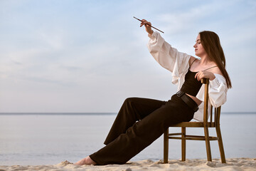 young female artist sits on wooden chair at beach, brush in hand, gazing calmly at horizon. Serene...