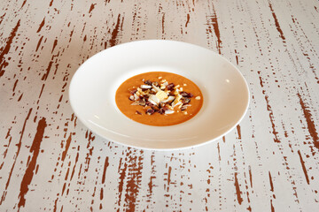 A white plate with a red soup on it. The soup has nuts in it. The plate is on a table