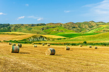 Fototapeta premium Scenic view at picturesque golden sunset valley in a green shiny field with hay stacks, bright cloudy sky , golden sun rays and beautiful hills on background, summer valley landscape