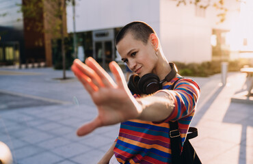 Stylish buzzcut woman in striped shirt blocks camera playfully with hand, smiling confidently...