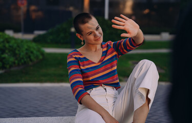 Close-up of young woman smiling softly while raising hand to block sun rays, sitting in urban park...